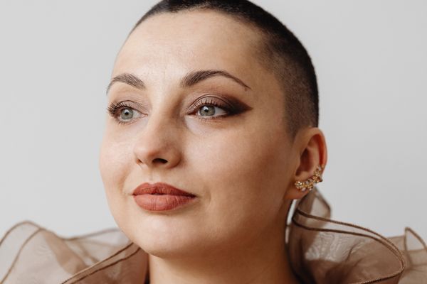 Microneedling 1 A close-up portrait of a person with a shaved head and fair skin looking off-camera. They are wearing bold, winged eyeshadow, matte lipstick, and a decorative gold ear cuff. The image showcases smooth skin texture and refined facial contours.