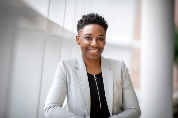 Microneedling 3 woman with short, styled hair. She is wearing a grey blazer over a black top and standing in a brightly lit, modern environment with soft-focus background pillars. Her skin appears vibrant, even-toned, and firm.
