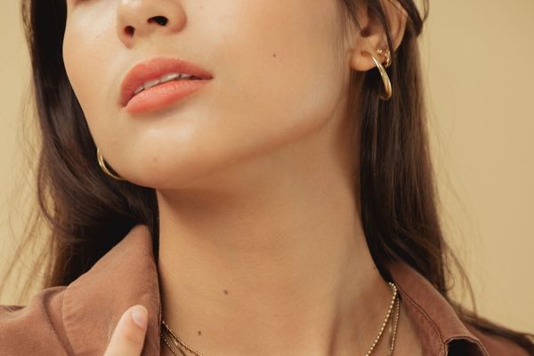 Microneedling 4 A close-up shot of a young woman's lower face, jawline, and neck. Her skin is exceptionally smooth, clear, and glowing, demonstrating a "post-treatment" or healthy skin result. She is wearing gold hoop earrings and a brown collared shirt.
