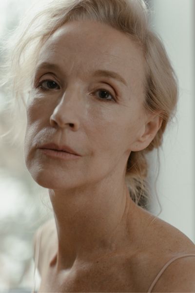 fine lines A portrait of a mature woman with fair skin and blonde hair. The lighting emphasizes natural aging features, including fine lines around the eyes, forehead, and mouth, as well as slight skin folding on the neck.