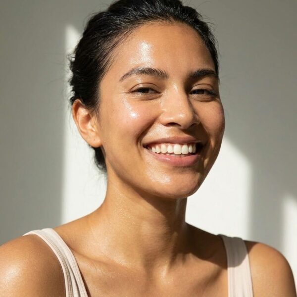 A vibrant photograph of a smiling woman with visibly hydrated, dewy skin standing outdoors in soft daylight.