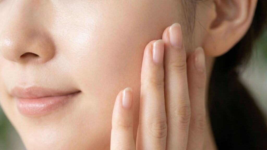 A soft, natural close-up photograph capturing a woman's manicured hand gently resting against her very smooth, glowing cheek.